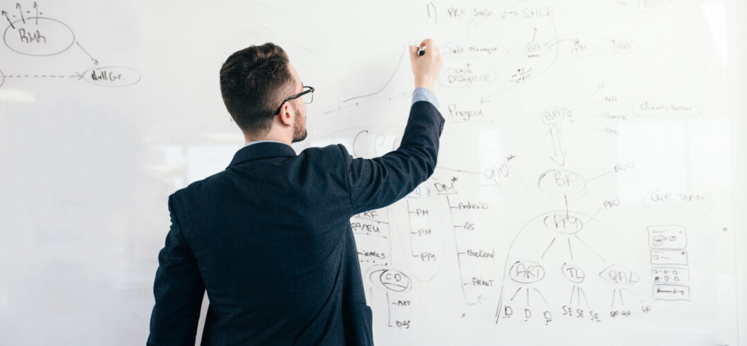 Young attractive dark-haired man in glasses is writing a business plan on whiteboard. He wears blue shirt and dark jacket. View from back.